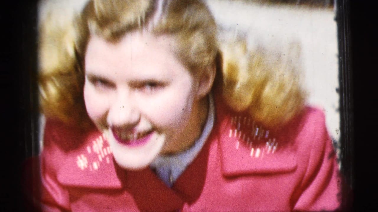Blonde woman in a red coat leans toward the camera outdoors.