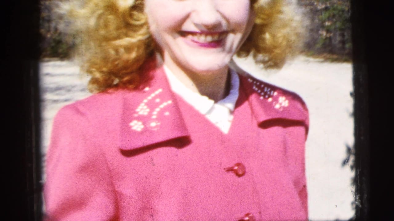 Smiling woman in a red coat is partly cropped in an outdoor frame.