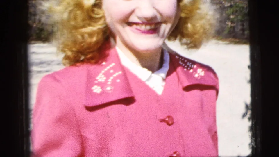 The woman in the red coat smiles in bright outdoor light, with her face partly cropped by the home-movie framing.
