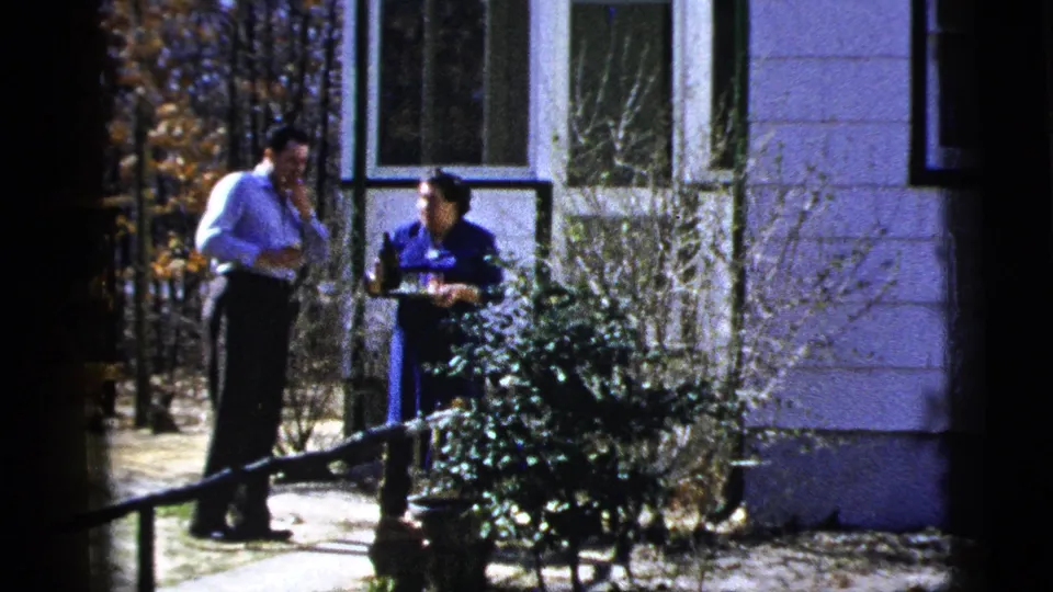 Two adults appear to converse outside a residence, with shrubs, steps, windows and trees visible in the 1956 New York home-movie frame.