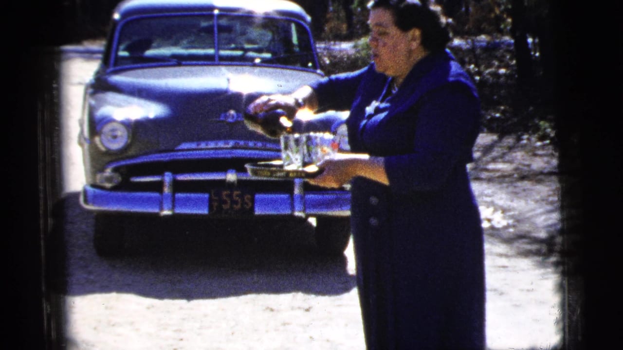 A woman in a dark blue dress serves beverages from a tray outdoors, with a 1950s sedan visible behind her.