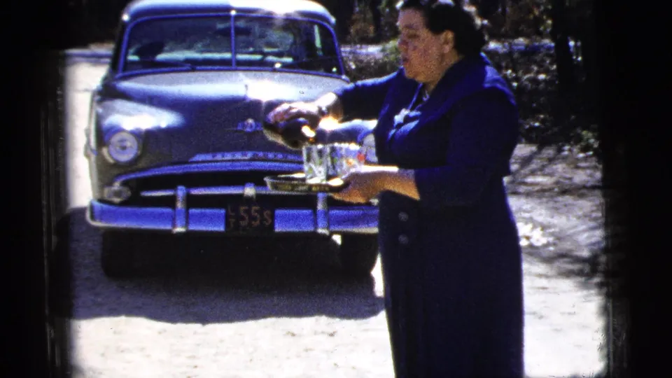 A woman in a dark blue dress serves beverages from a tray outdoors, with a 1950s sedan visible behind her.