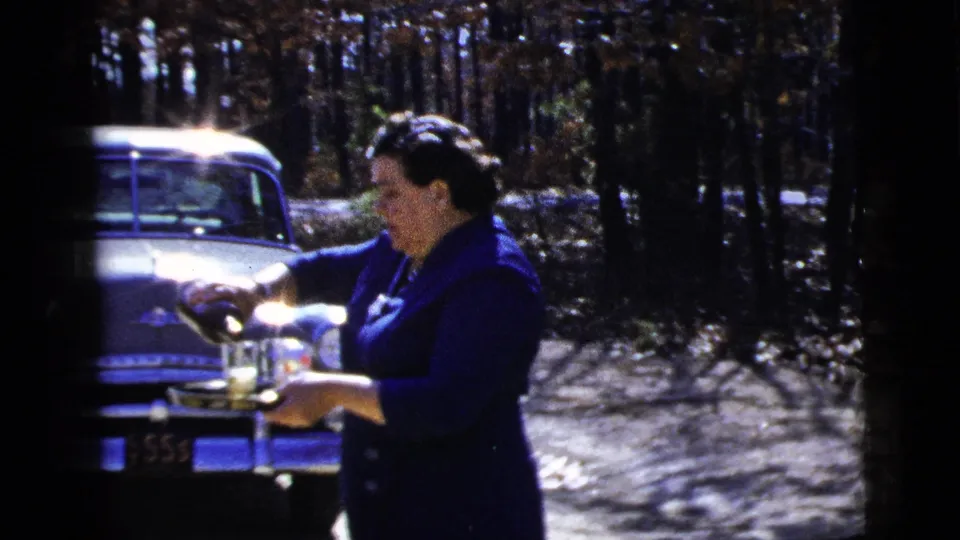 The woman continues serving beverages outdoors, partly framed by film edge and the sedan behind her.