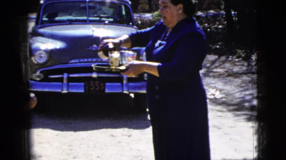 A woman in a blue dress holds a tray of sparkling beverages near a parked 1950s sedan in the outdoor scene.