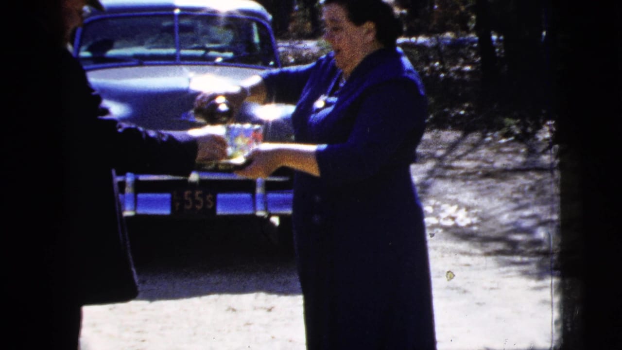 Woman in a dark blue dress holds a tray of glasses near a parked classic car.
