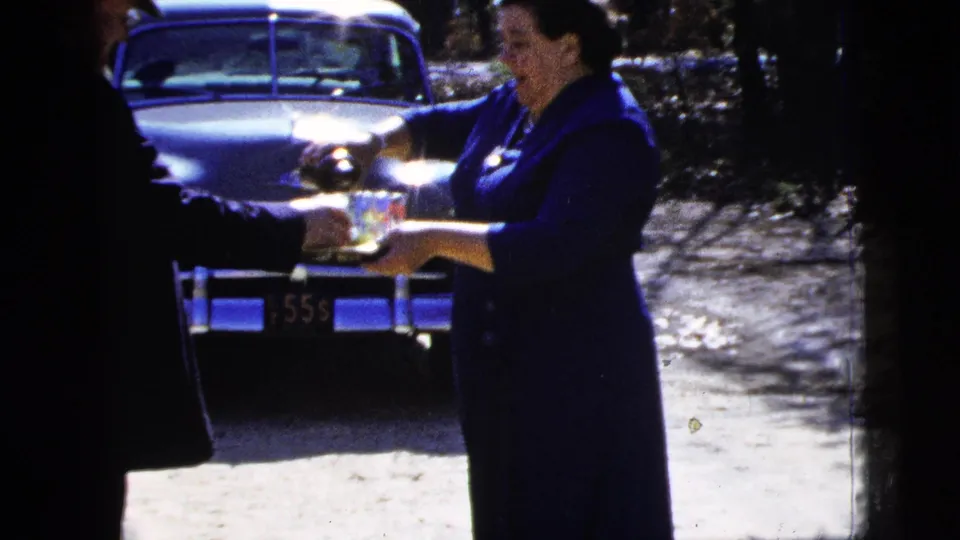 In 1956 New York City home-movie footage, a woman in a dark blue dress serves drinks from a tray beside a parked classic car.