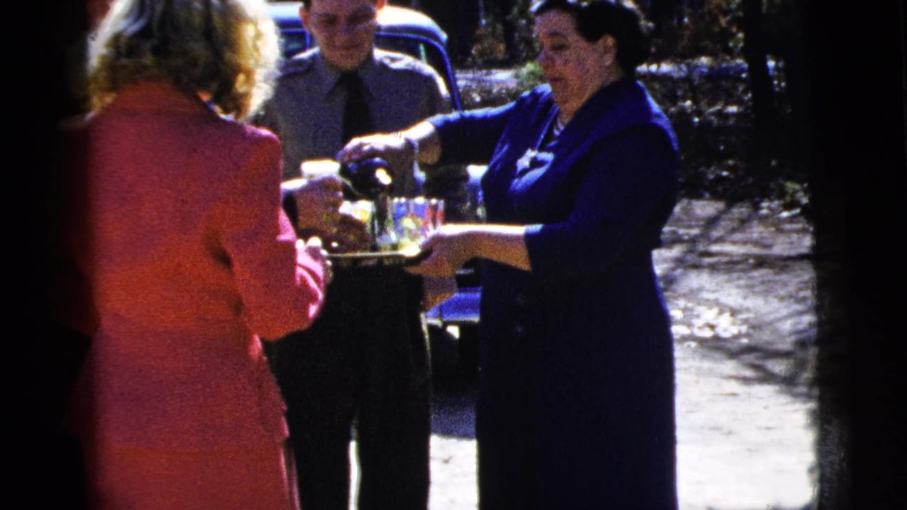 Woman pours from a bottle toward glasses on a tray as two adults stand nearby.