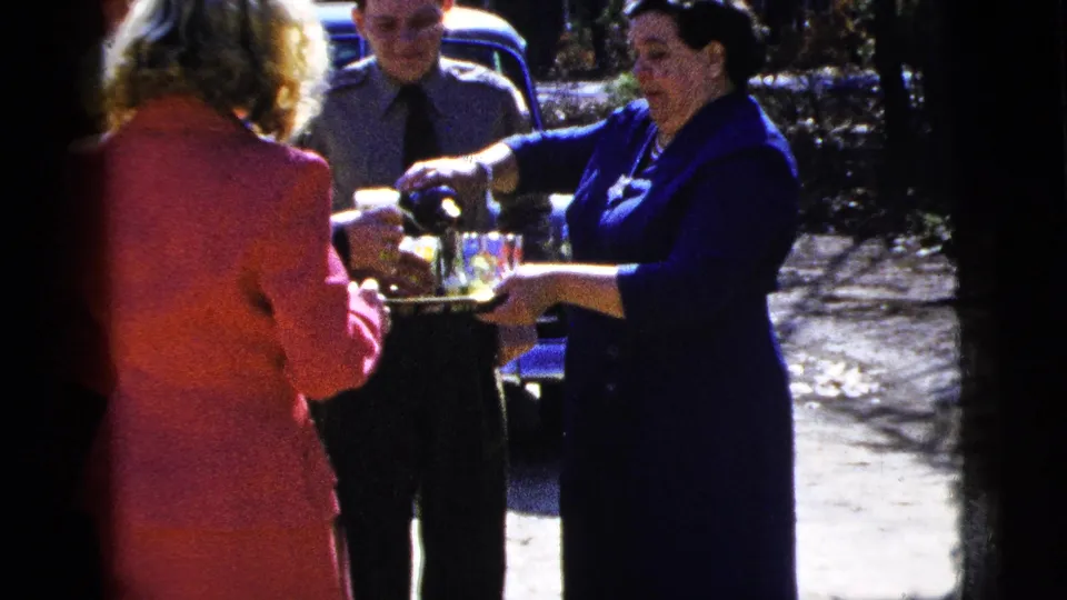 A woman in a dark blue dress pours drinks for adults near a classic car in the supplied 1956 New York City setting.