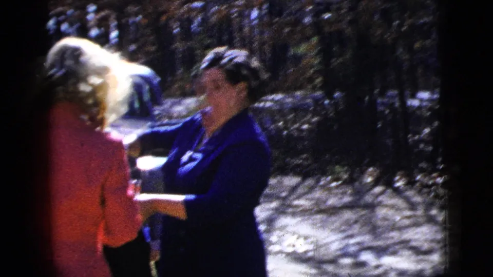 The home-movie frame shows two women at an outdoor gathering, with a tray of drinks held between them in bright sunlight.