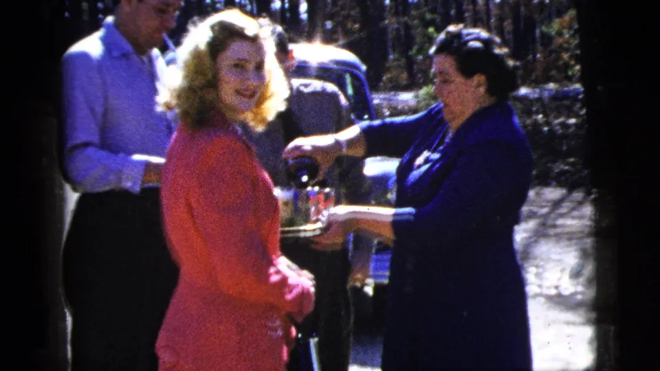 A woman in a pink suit looks toward the camera as another woman pours drinks from a tray near a parked classic car.