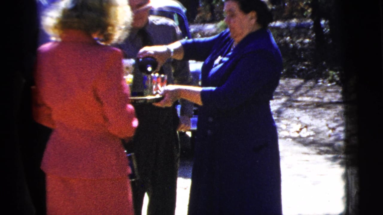 1956 New York City home-movie frame showing adults gathered outside while a woman in a dark blue dress pours drinks from a silver tray.