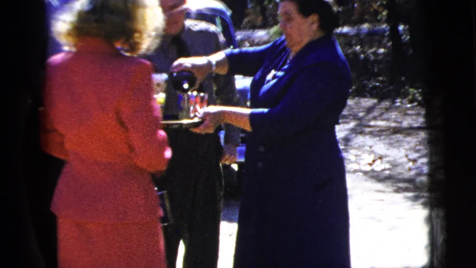 1956 New York City home-movie frame showing adults gathered outside while a woman in a dark blue dress pours drinks from a silver tray.