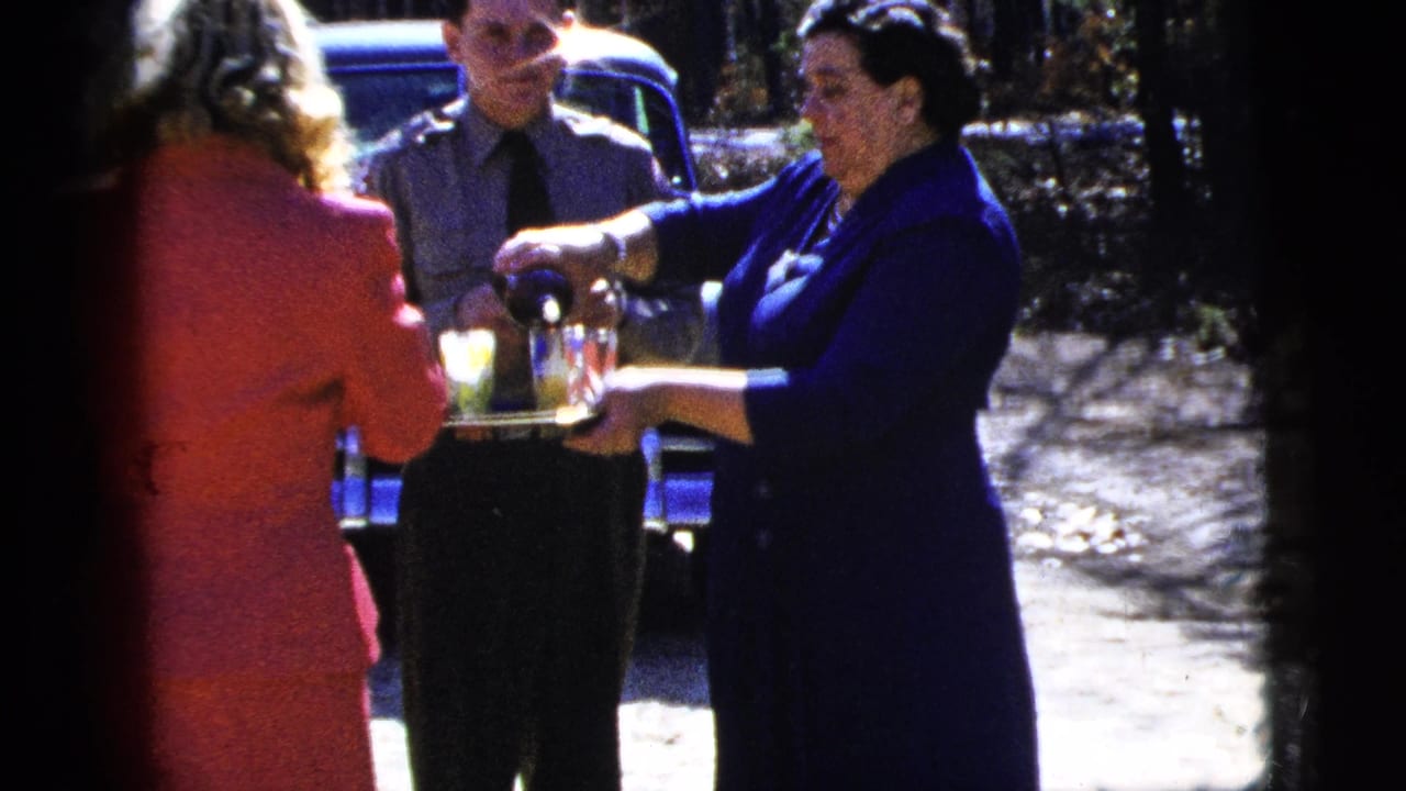 Adults stand outdoors in New York City as a woman pours drinks, with part of a classic car visible behind them.