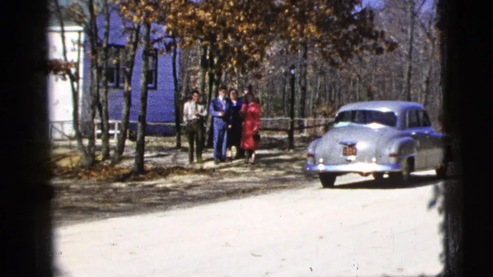 1956 New York City home-movie view of adults standing near a tree-lined road with a pale rounded classic car parked nearby.