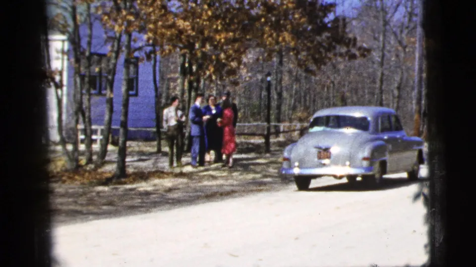Wide 1956 New York City home-movie frame with adults grouped near trees and a pale mid-century car parked on the road.