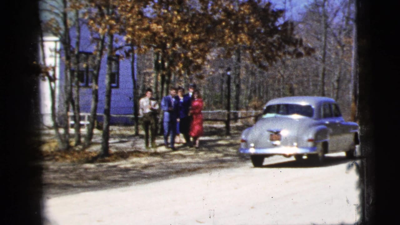 Group of young people near a silver sedan on a dirt road beside a blue building and trees.