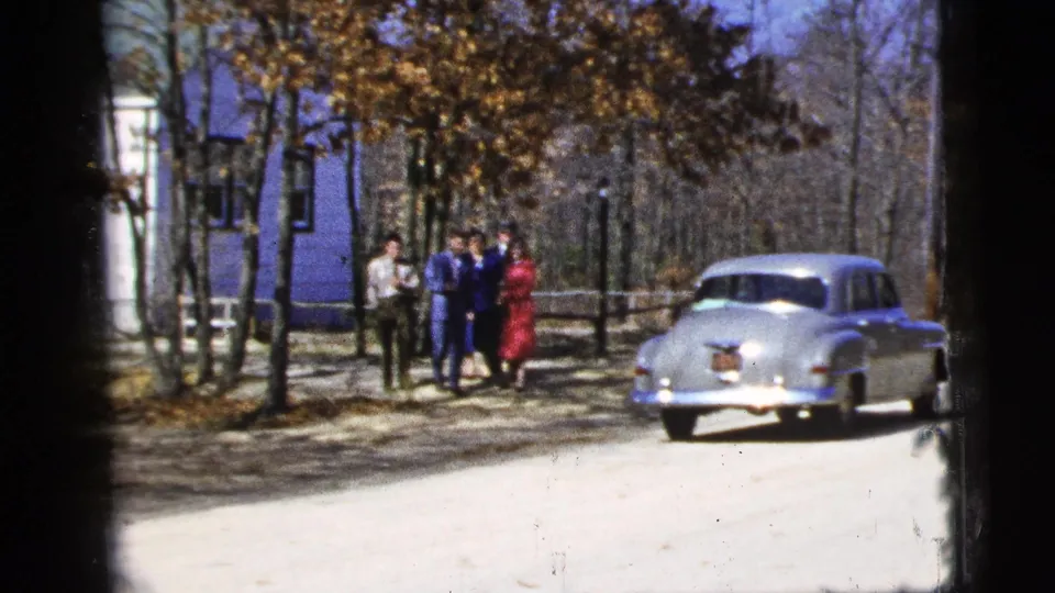 1956 8mm frame of young people standing near a silver sedan on a dirt road in a wooded New York City setting.