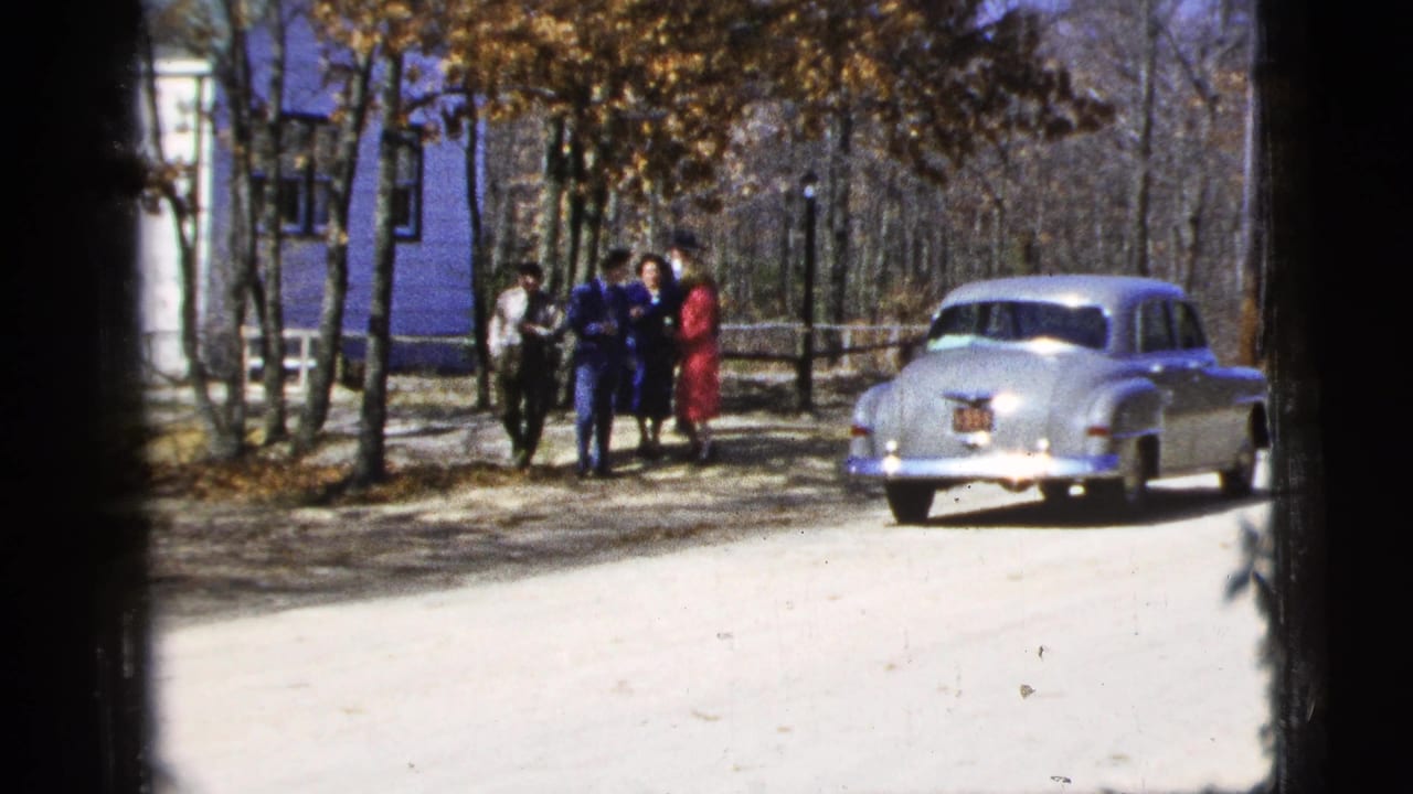 Five young people stand near a silver sedan on a pale dirt road with trees behind them.