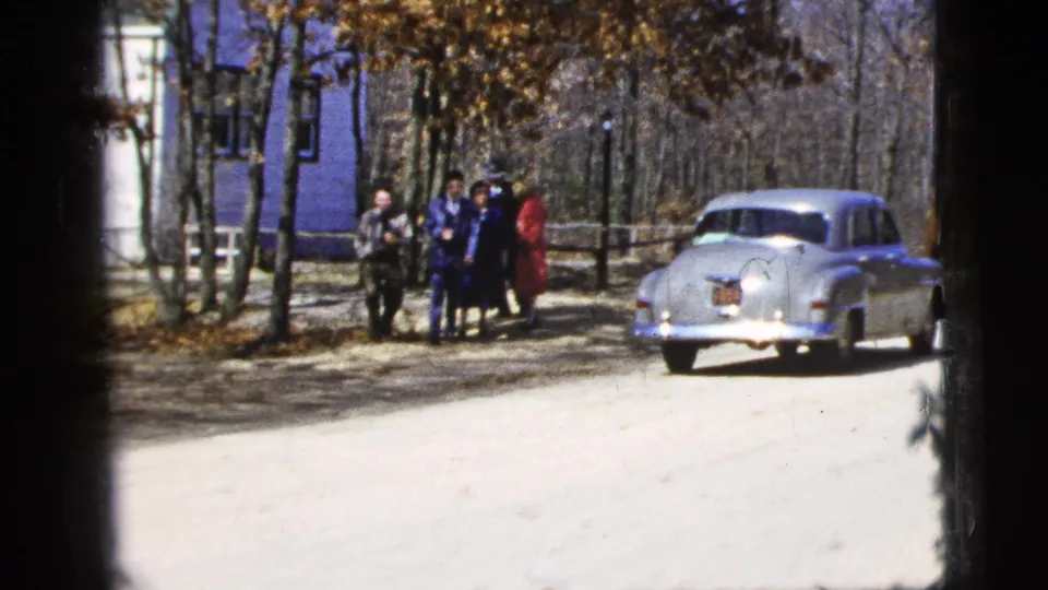 In a 1956 home-movie frame, the group shifts position near the silver sedan, with a blue building and bare trees in the background.