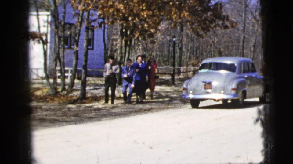 Final 1956 8mm frame shows young people walking on a dirt road near a silver sedan in a wooded New York City setting.