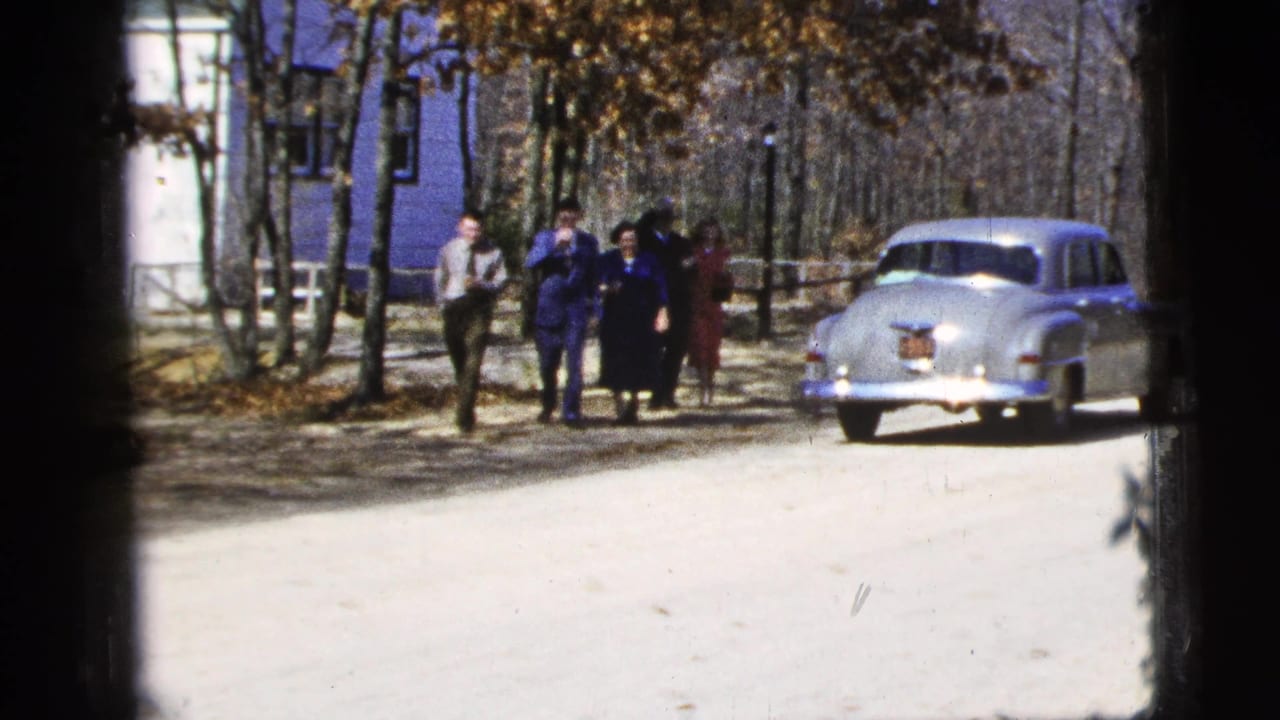 Five adults walk on a pale dirt road near a parked silver sedan and blue building.