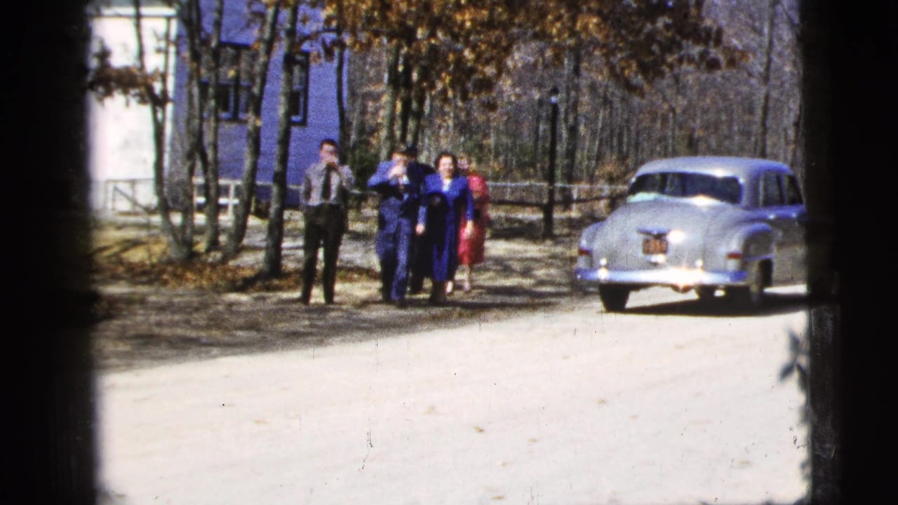 A group of adults stands and begins walking near a silver sedan on a wooded road.