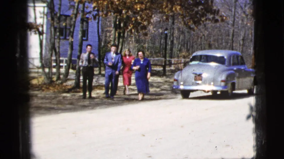 Five adults in suits and dresses walk toward the camera on a dirt road, with a mid-century silver sedan behind them in the 1956 New York clip.