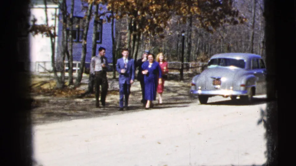 Near the end of the clip, five adults continue toward the camera on a tree-lined dirt road with a silver sedan parked to their right.