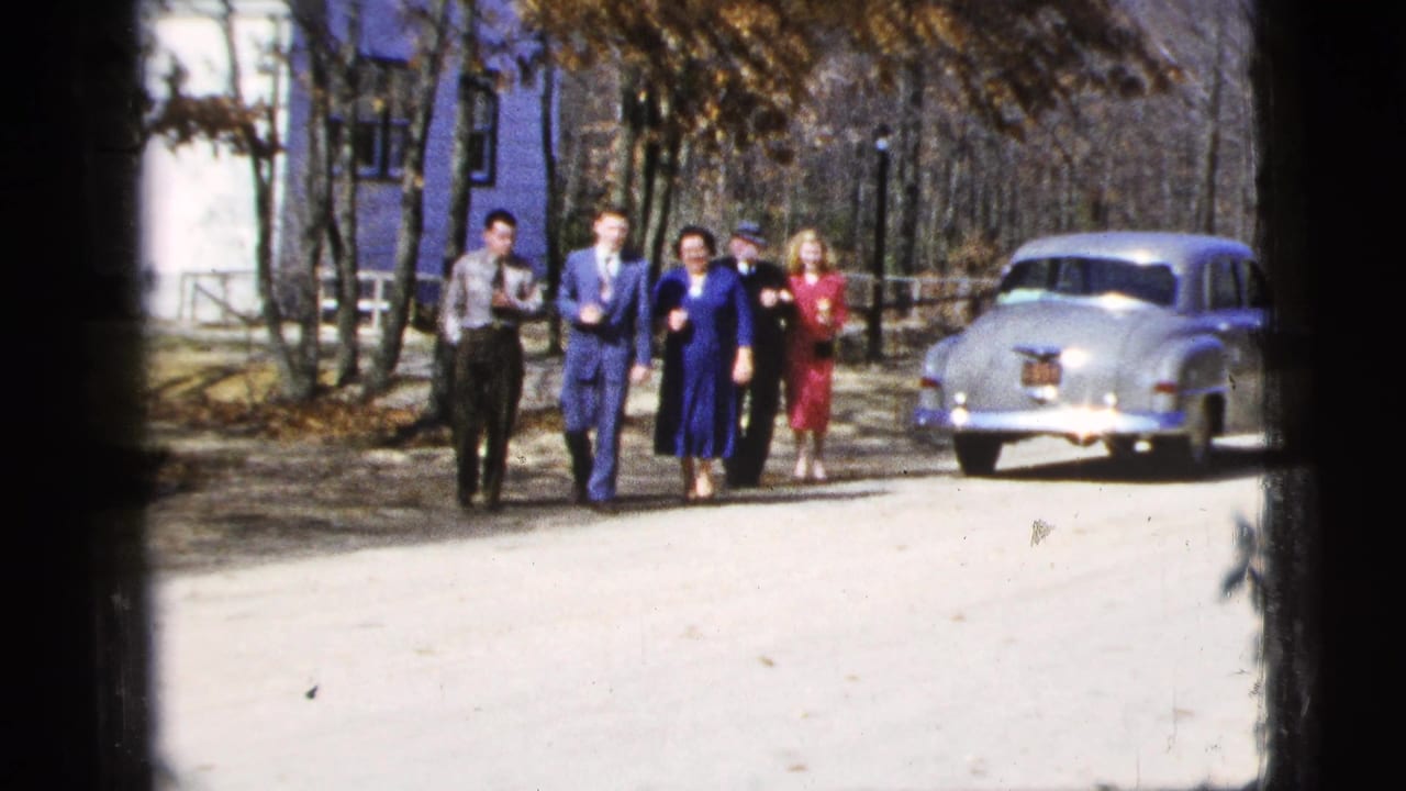 Five adults walk on a dirt road near a light-colored sedan and a blue-and-white building.