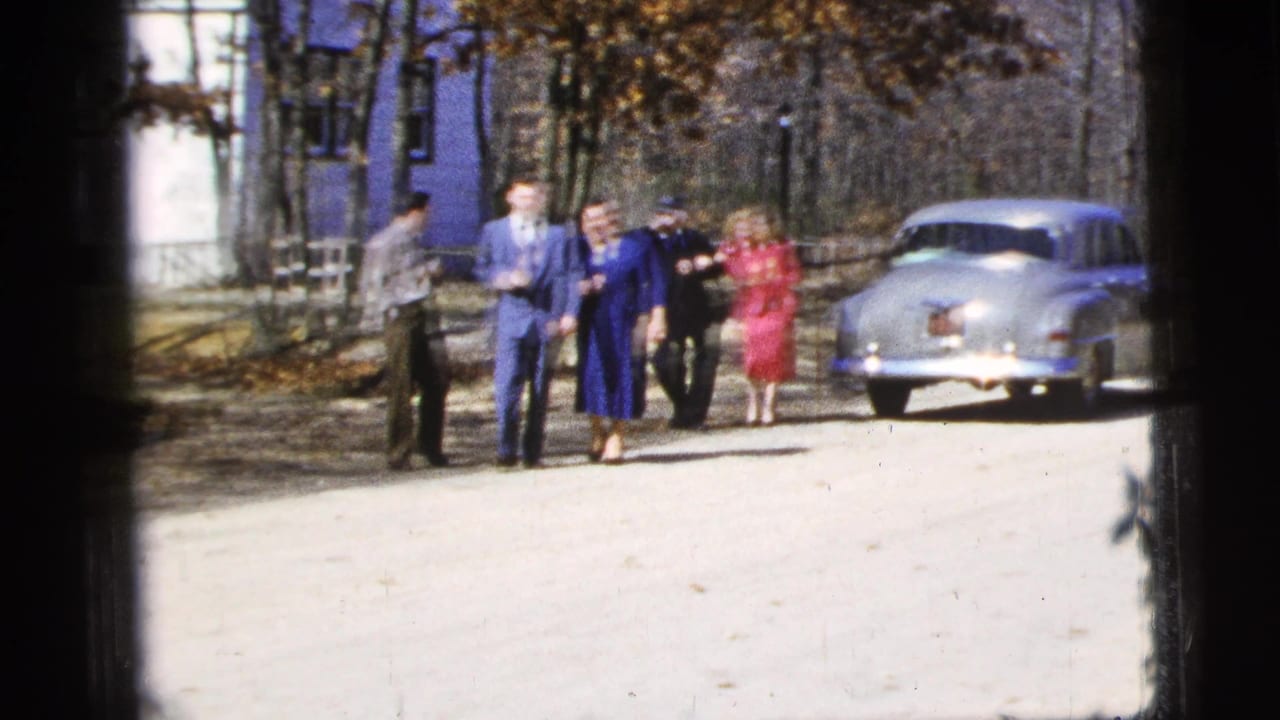 The group advances along the bright dirt road, with period clothing, trees, and a light-colored classic sedan visible in the background.