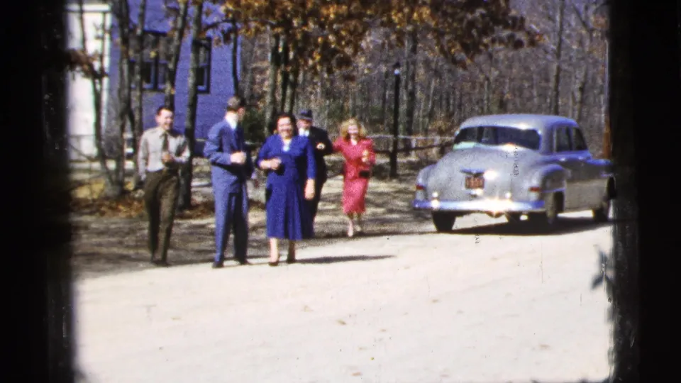 Three adults are most prominent as the group nears the camera, with the silver-toned sedan, trees, and building still visible behind them.