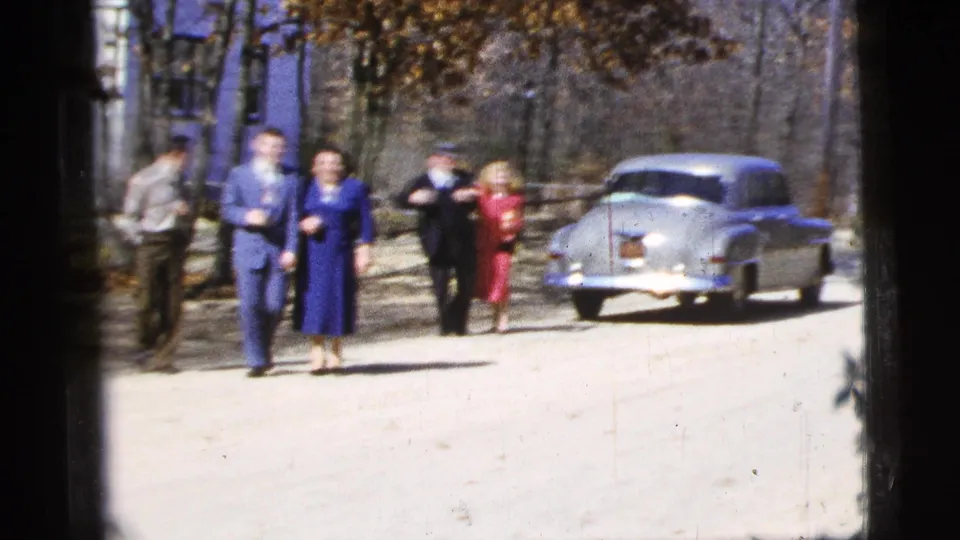 The final frame shows adults in period dress moving down the road, with the parked sedan and wooded background anchoring the 1956 home-movie setting.