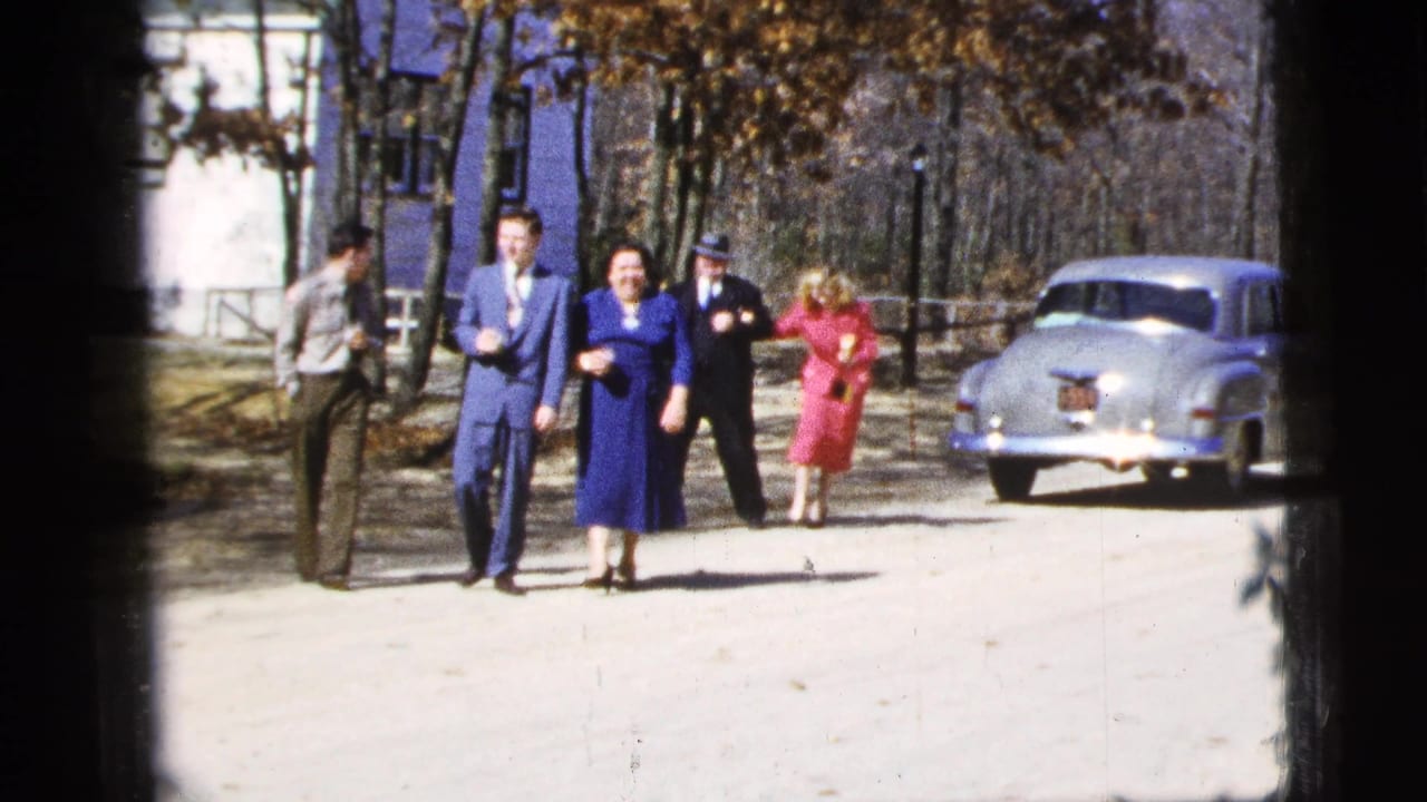 Adults in formal clothes walk along a sunlit street near a pale vintage sedan.