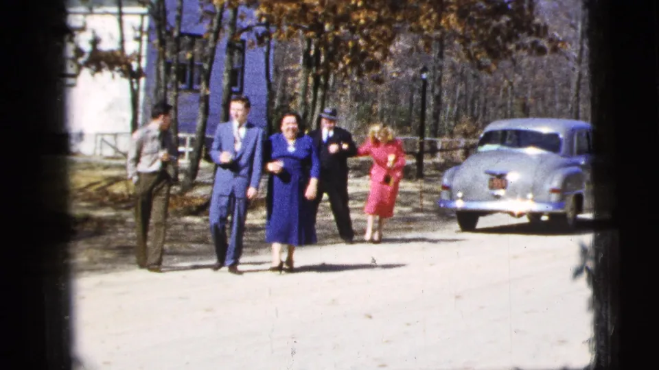 1956 New York City home-movie frame showing well-dressed adults walking toward the camera near a rounded pale sedan.