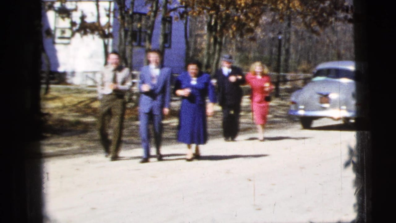 A group of adults walks toward the camera with a pale sedan parked to the right.