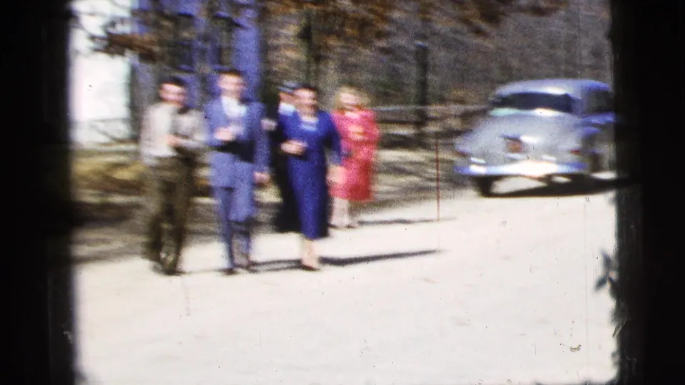 Soft 8mm frame of adults in formal mid-century clothing walking on a sunlit New York City street near a parked sedan.