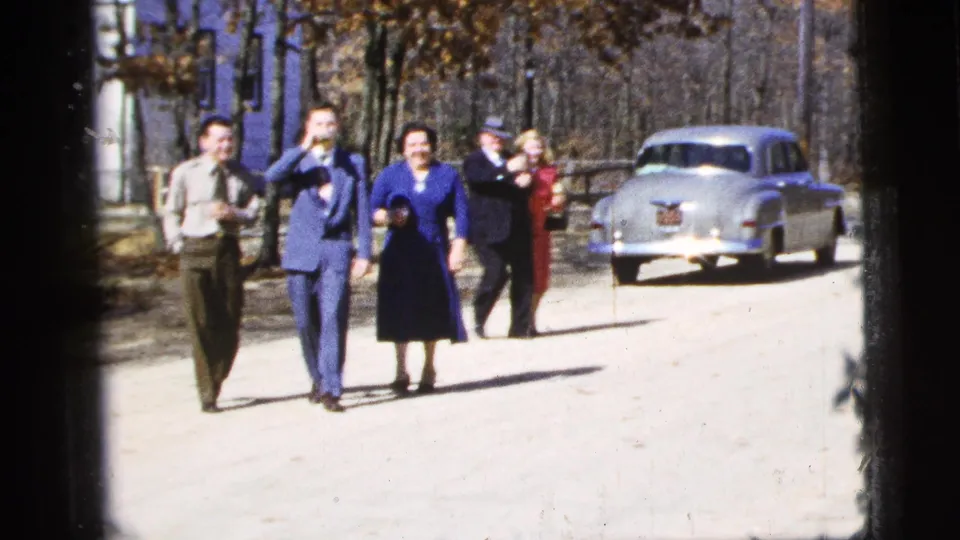 1956 New York City frame showing a couple walking arm-in-arm, followed by other formally dressed adults and a pale sedan.