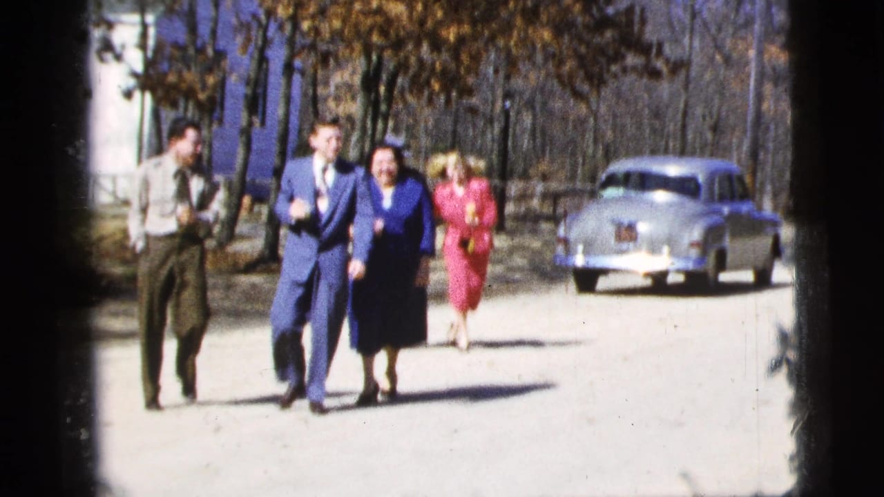 Well-dressed adults continue toward the camera on a New York City street, with a silver mid-century sedan parked behind them.