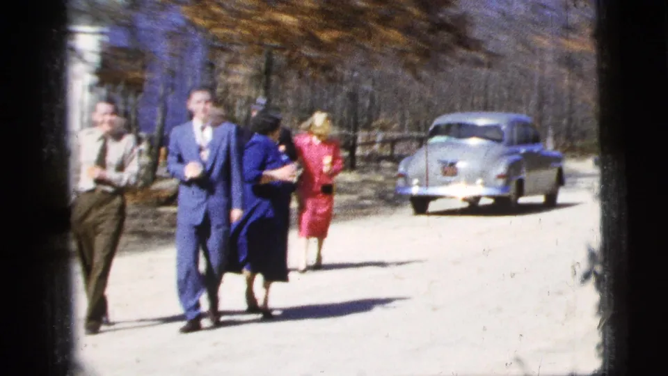 The group moves closer in the 1956 home-movie street scene, with suited men, a woman in blue, and a woman in pink visible.