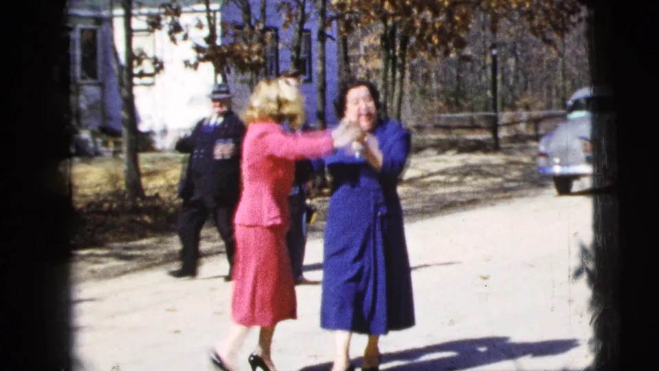 Two women in blue and pink outfits share a playful greeting or dance on the sunlit New York City street as others stand nearby.