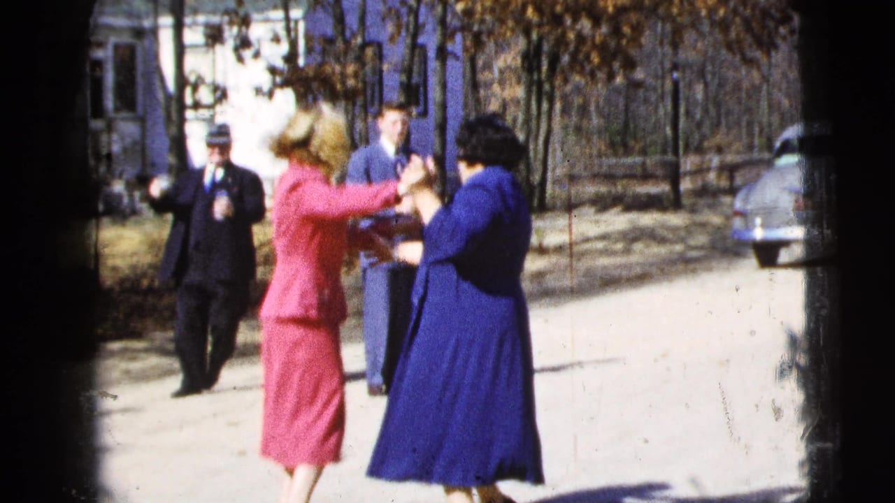 In 1956 New York City home-movie footage, two women in bright dresses dance on a street with adults, trees, and a parked car behind them.