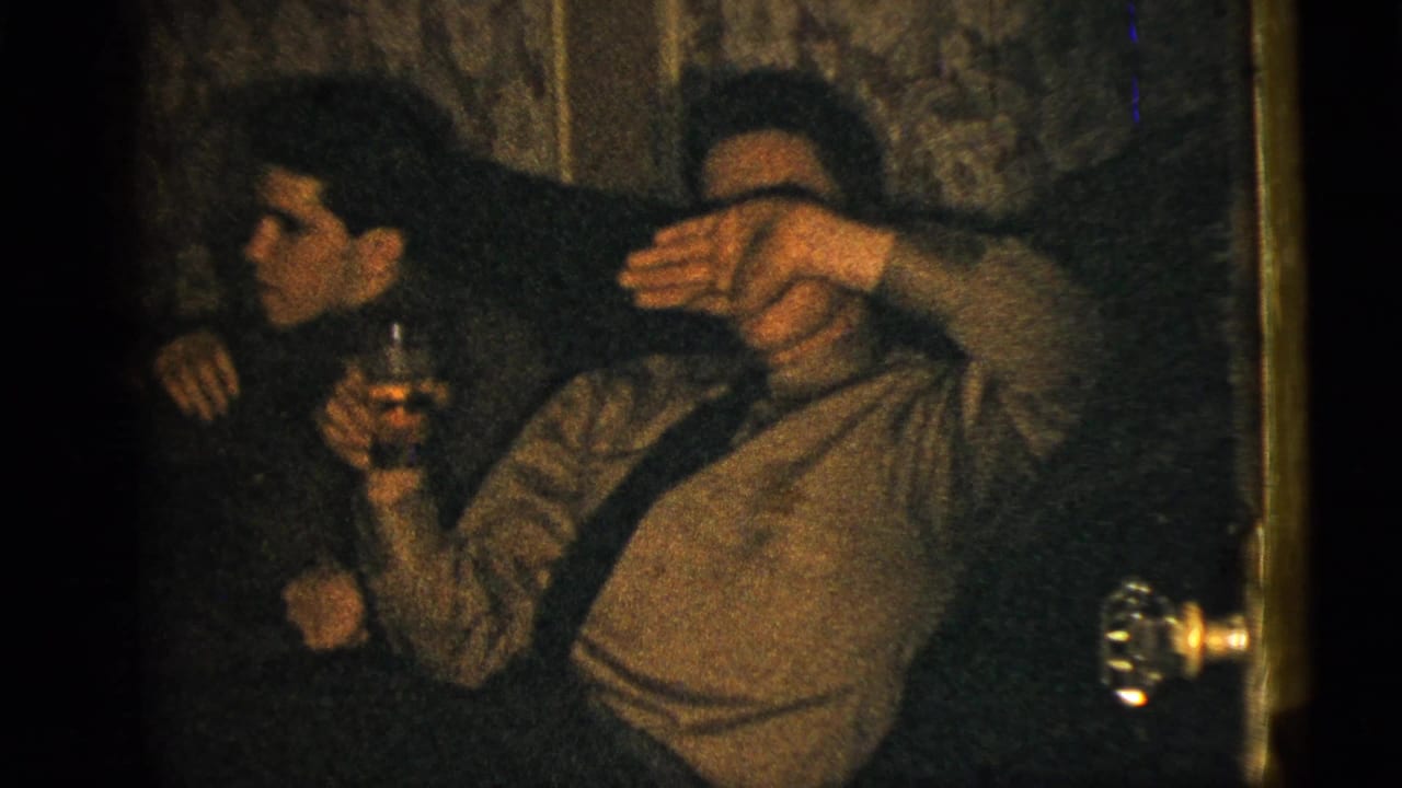 Dim interior 8mm frame from the 1956 New York City clip showing two seated men, one with a drink and another shielding part of his face.