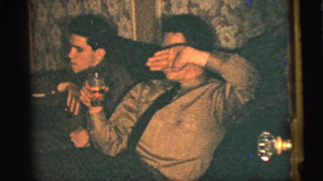Warm, grainy 8mm interior frame showing two men seated closely, one holding a drink while the other raises an arm across his face.