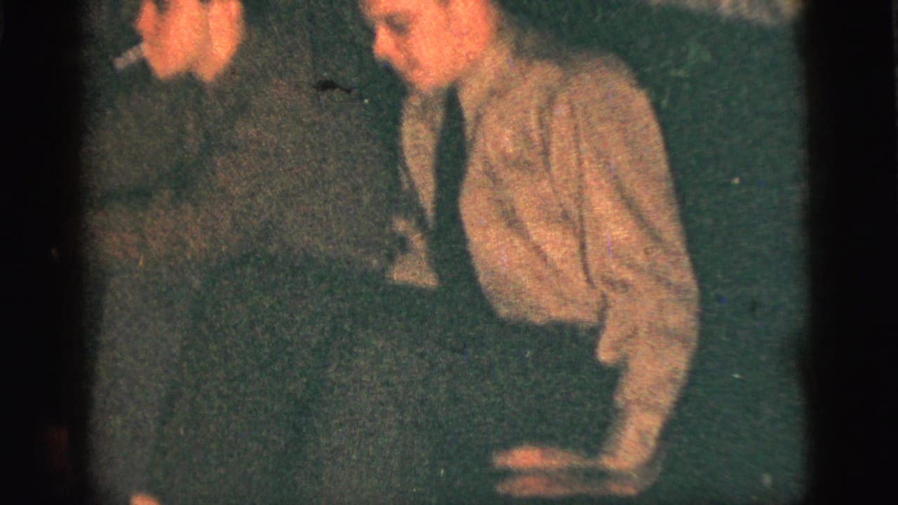 Dim 1956 New York City home-movie frame showing two men seated indoors, one smoking while the other leans close in soft focus.