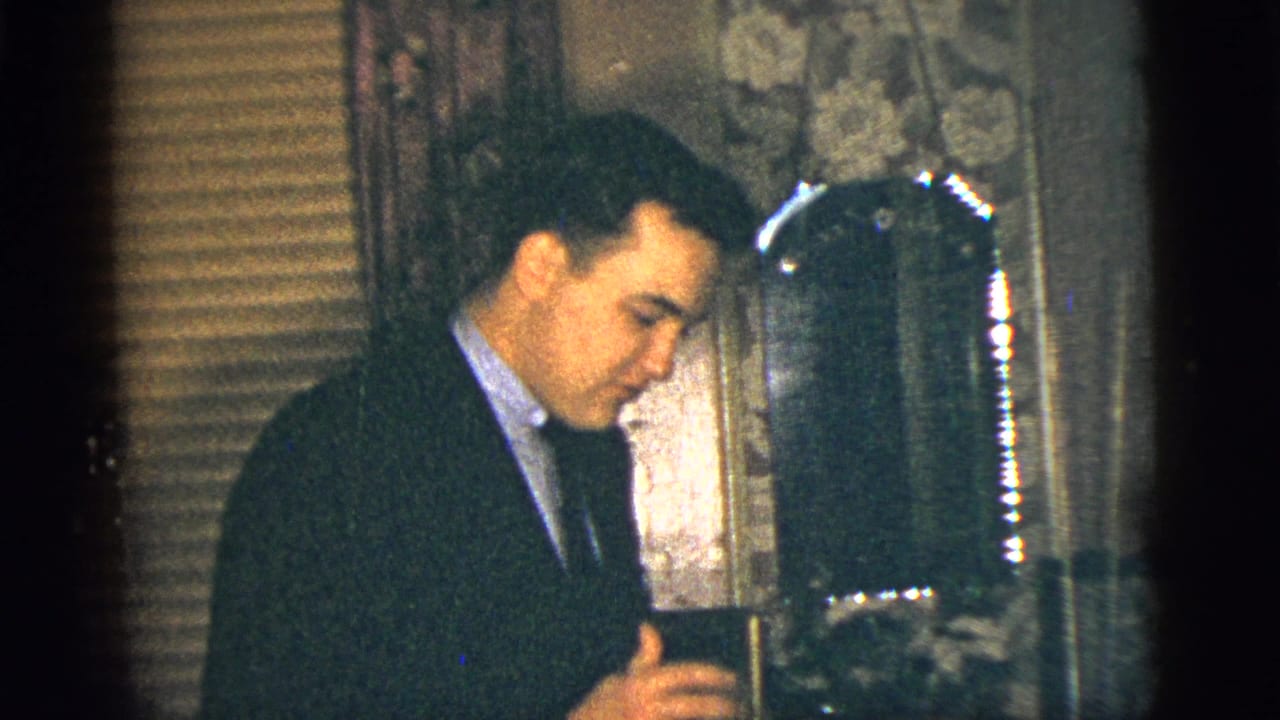 In 1956 New York City home-movie footage, a suited man leans beside a mirror in a dim private room with wallpaper and blinds.