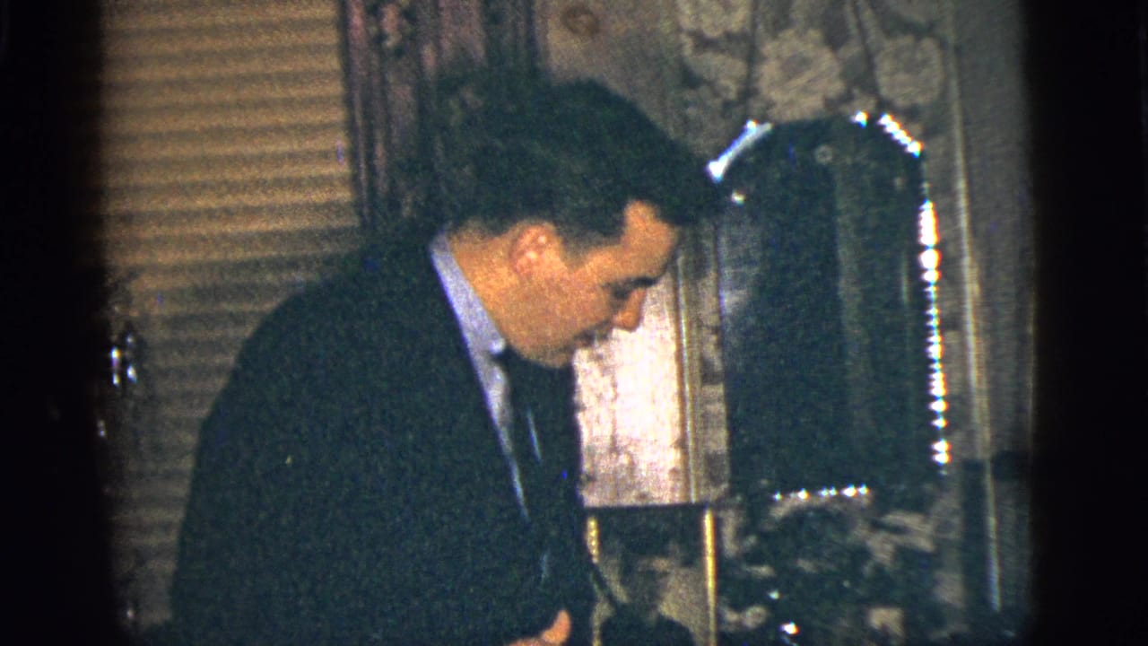 Man in a dark suit looks downward beside a mirror and household decor.
