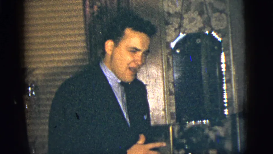 The 1956 home-movie frame shows a suited man near a mirror, with patterned wallpaper and blinds behind him in a private New York City interior.