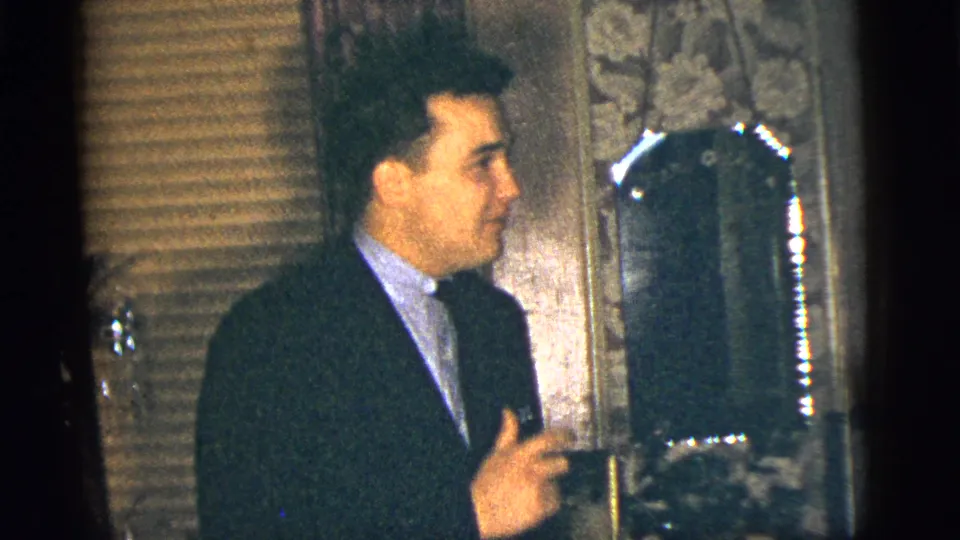 A suited man gestures near his chest while facing a mirror in a dim 1956 New York City private interior.