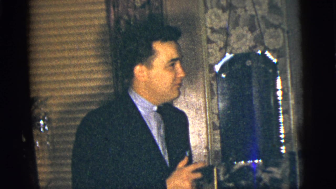 Man in a dark suit and tie stands beside patterned wallpaper and window blinds.