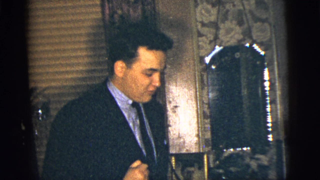 The man in a suit and tie looks down in a dim New York City interior, with blinds and patterned wallpaper behind him.